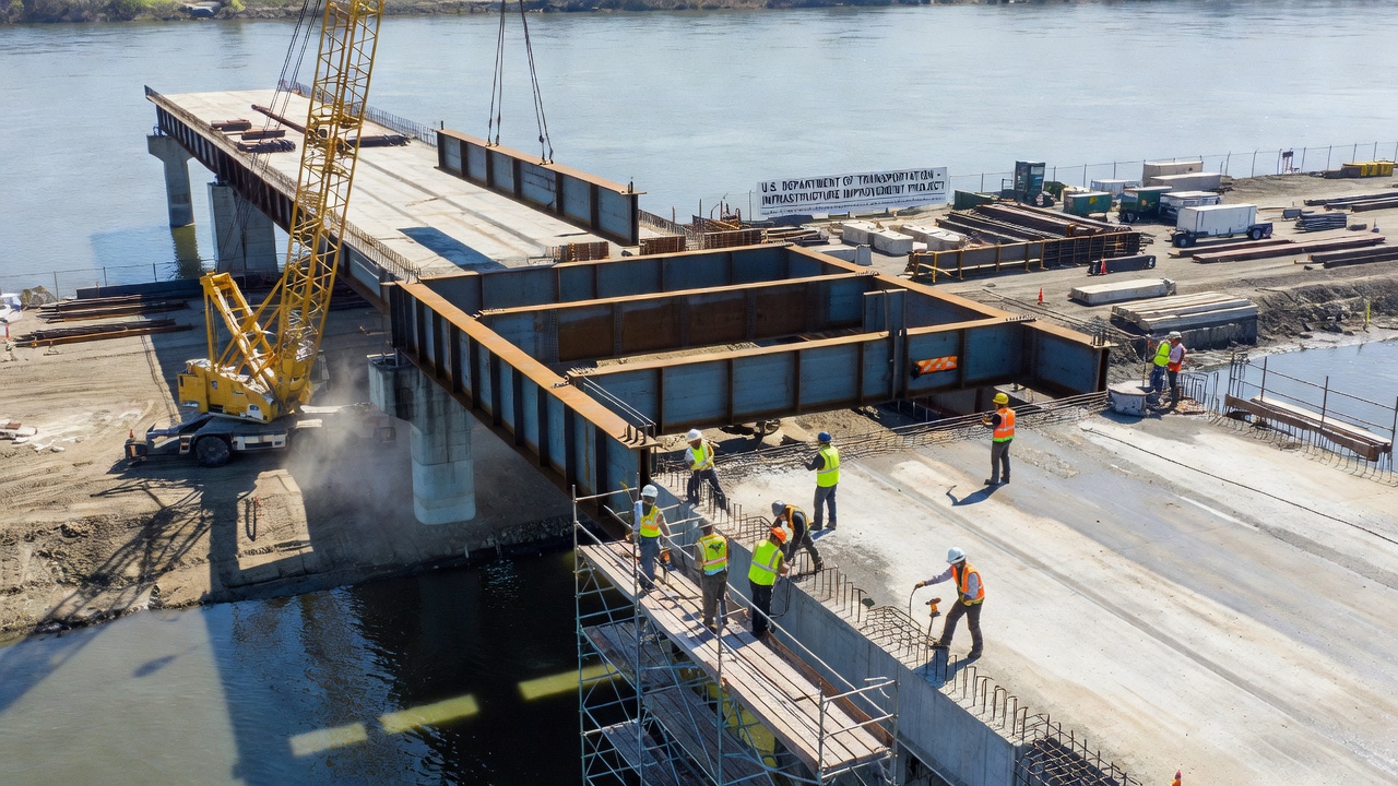 Chantier de construction d'un pont montrant un pont à poutres en acier avec tablier en béton en cours de construction, ouvriers et grue sur le site