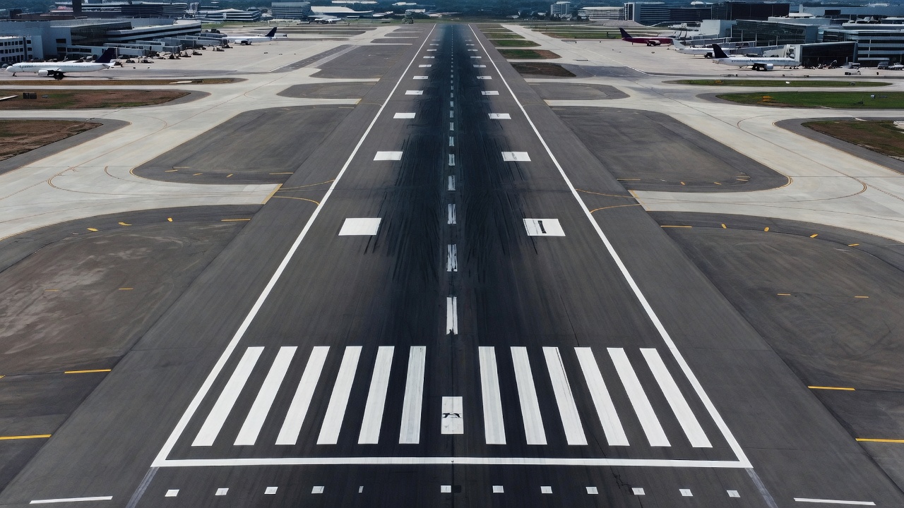 Aerial view of a commercial airport runway with clear white centerline markings and dashed stripes, precision instrument runway markings visible in daytime lighting